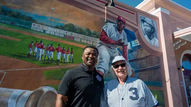 The man in the black polo and the man in the vintage pinstriped number 3 uniform pose together before the mural.