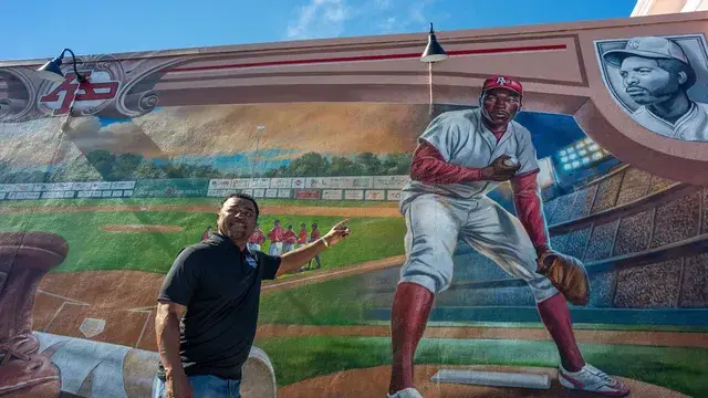 The man in black spreads his arms wide in front of the mural.