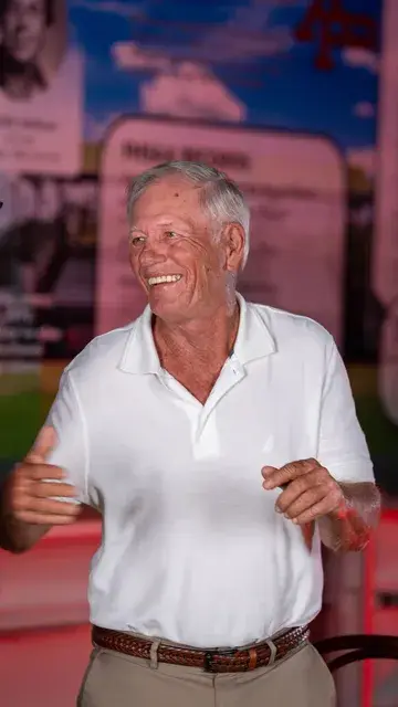 An older man in a white polo laughs under red lighting in front of the FHSAA records display.