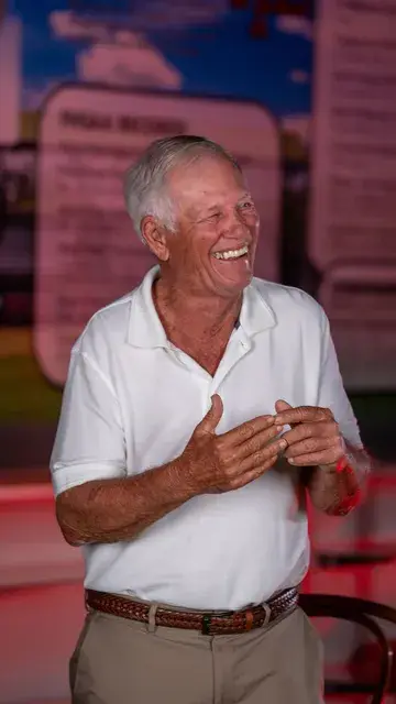 The man in the white polo, hands clasped, laughing in the red-lit gallery.
