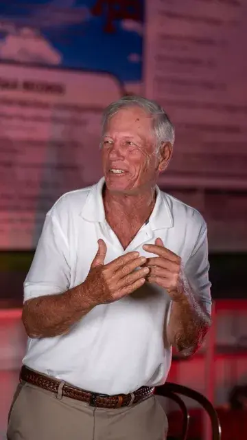 The man in the white polo gestures as he talks in the red-lit gallery.