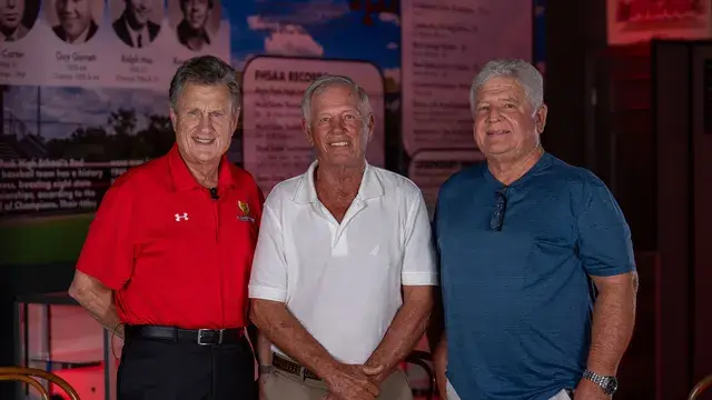 Three men pose in front of the records display, one in a red Devils polo.
