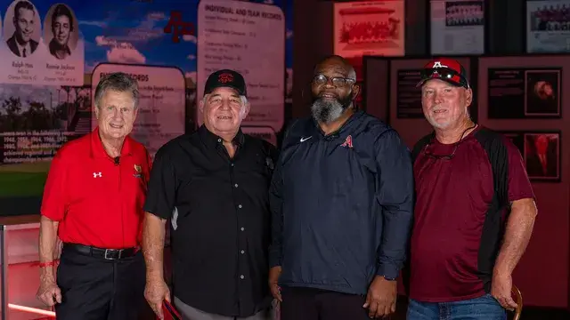 Four men pose before the records wall, two in red Devils polos and caps.