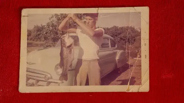 A faded snapshot of a young man holding up a fish beside a car.