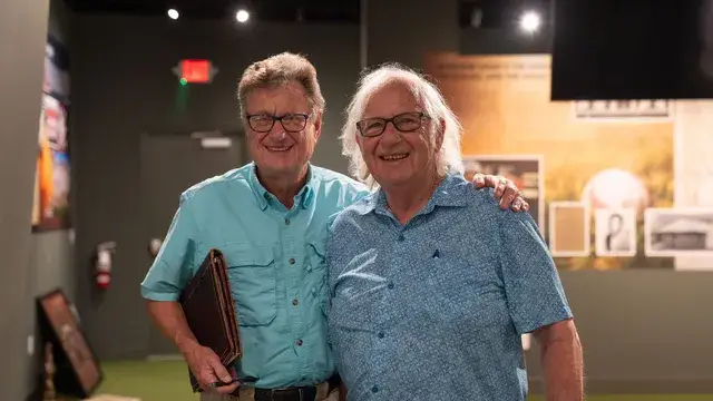 Two men pose in the gallery, one holding a folder.