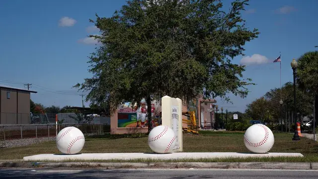 Giant baseball sculptures on a bat-shaped base beside the road, an oak tree behind.