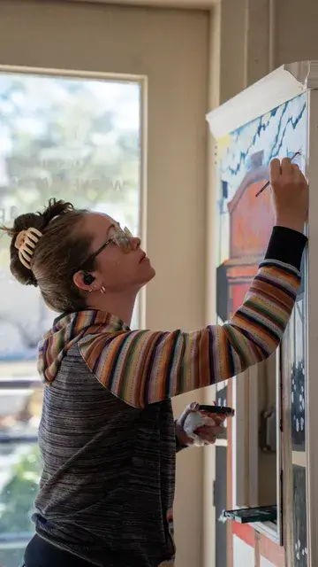 A woman paints a doorway mural by a window.
