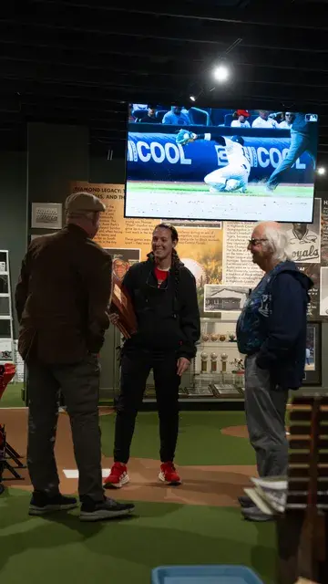 Three visitors talk beneath a screen showing a ball game.