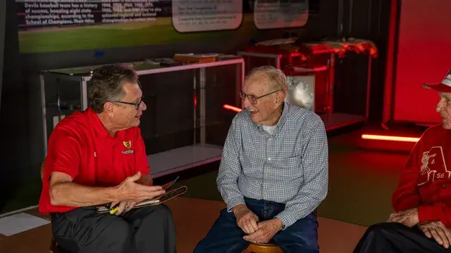 Three men talk in the red-lit gallery, one in a red polo, another in a checked shirt seated.