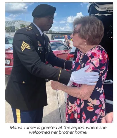 A soldier in dress uniform and white gloves clasps the hands of Marva Turner, an elderly woman in a floral dress, on the airport tarmac.